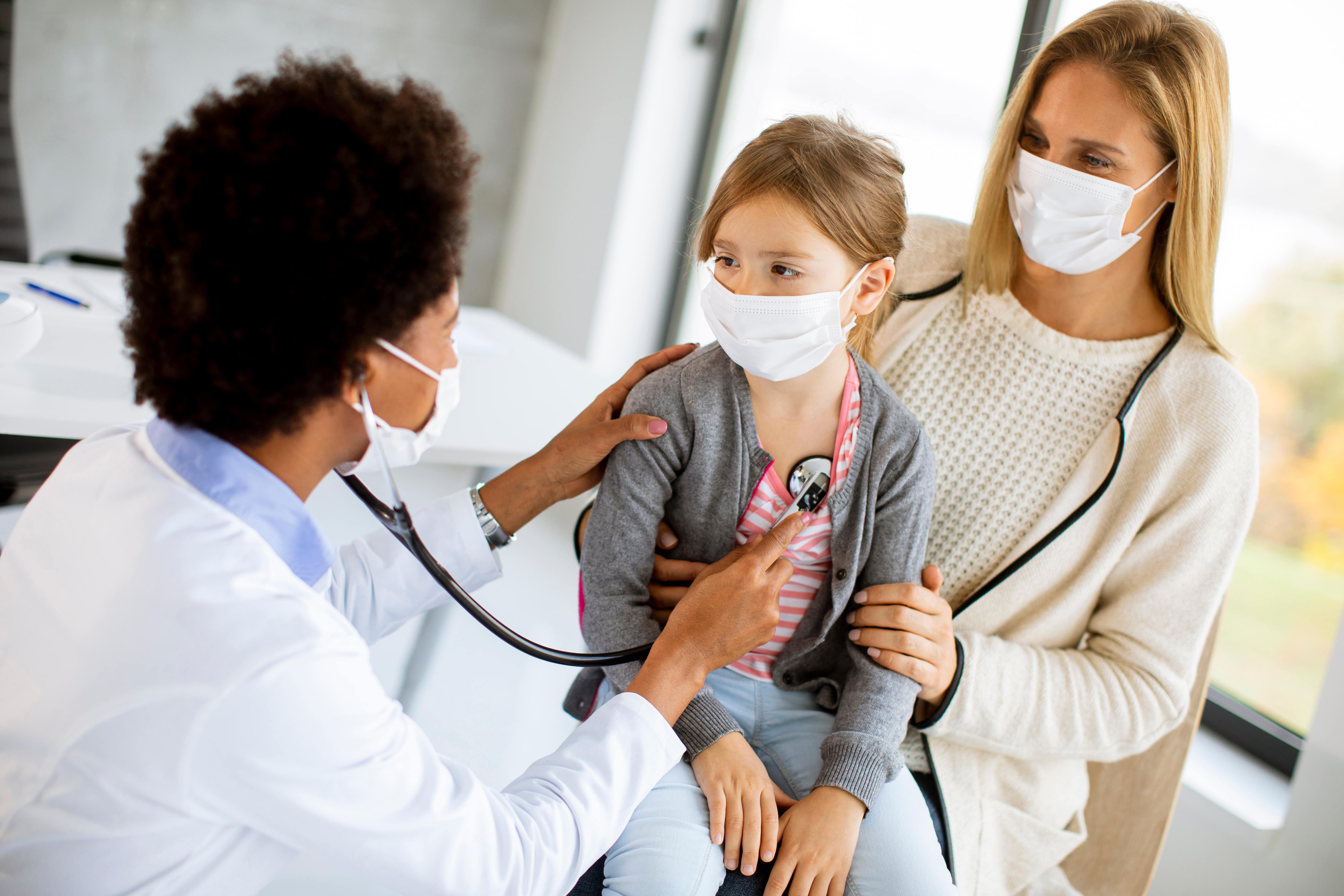 Masked doctor using a stethoscope on a young girl during an annual physical